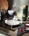 Round stainless steel grill pan by Alessi with ridged surface and squared handles, next to chopped radicchio.
