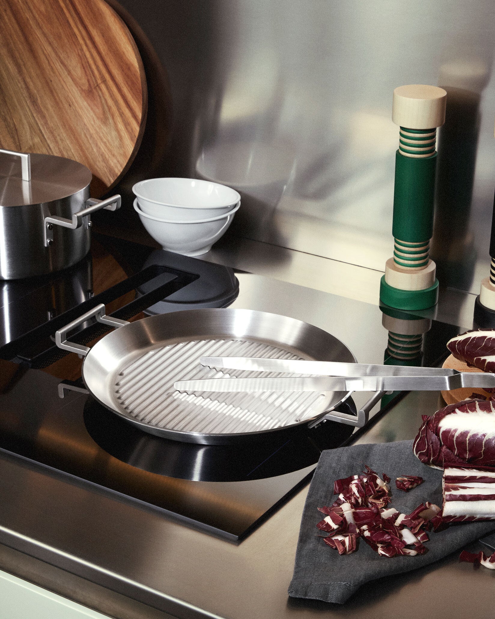 Round stainless steel grill pan by Alessi with ridged surface and squared handles, next to chopped radicchio.