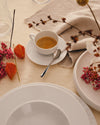 White ceramic espresso cup by Alessi on a saucer with a spoon, surrounded by fall decor on a beige tablecloth.