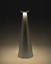 Gray cone-shaped Bedin table lamp by Alessi with a rounded top, emitting warm downward light onto a surface.