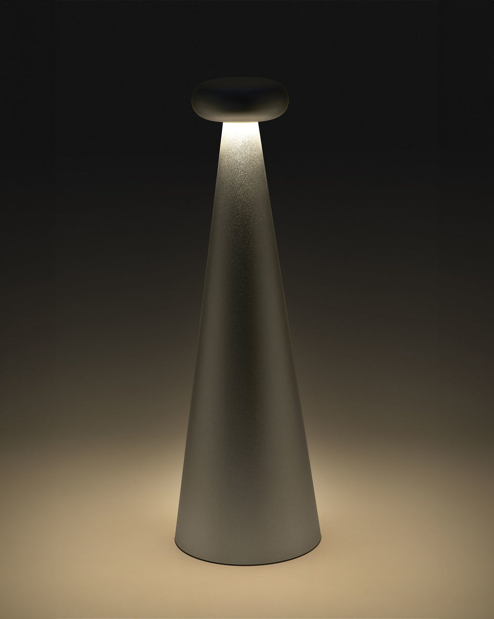 Gray cone-shaped Bedin table lamp by Alessi with a rounded top, emitting warm downward light onto a surface.
