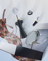Black and silver wine accessory set by Alessi, featuring a corkscrew, stopper, and drip ring with a smiling face design, floating above an open box.