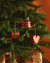 Three red and green Christmas tree ornaments by Alessi: a house, geometric heart, and space gift, on a green tree.