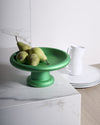 Vibrant green fruit bowl by Alessi, a wide, shallow design on a pedestal, holding four green pears on a marble counter.