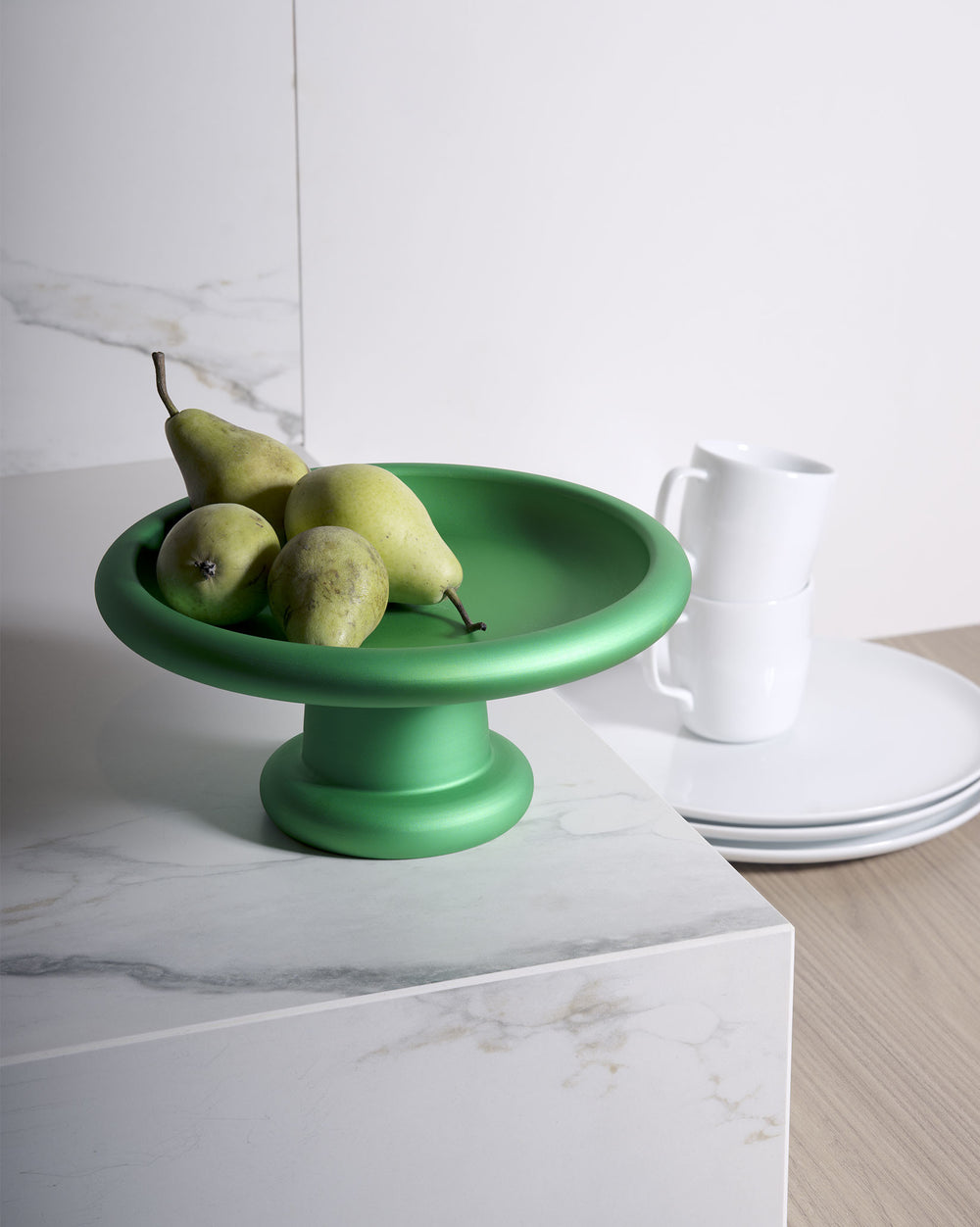 Vibrant green fruit bowl by Alessi, a wide, shallow design on a pedestal, holding four green pears on a marble counter.