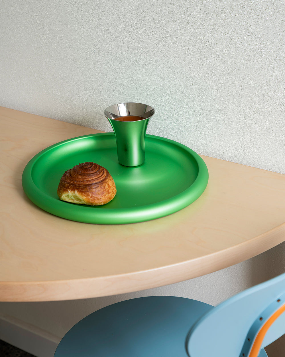 Green round metallic tray by Alessi, holding a matching cup and a pastry on a wooden table.