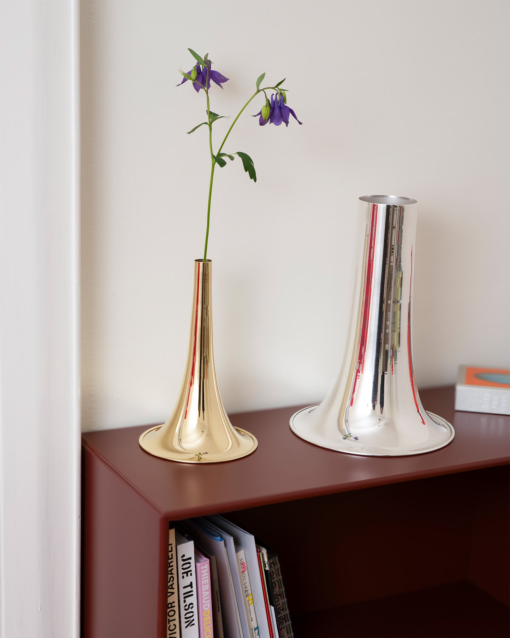 Gold trumpet vase by Alessi with a purple flower, next to a silver version on a brown shelf.
