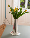 Silver trumpet vase by Alessi with polished metal finish, vertical etched lines, and red reflections, holding yellow tulips on a grey table.