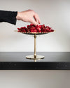 Gold and silver centerpiece stand by Alessi with a scalloped edge, filled with red-wrapped candies, a hand reaching.