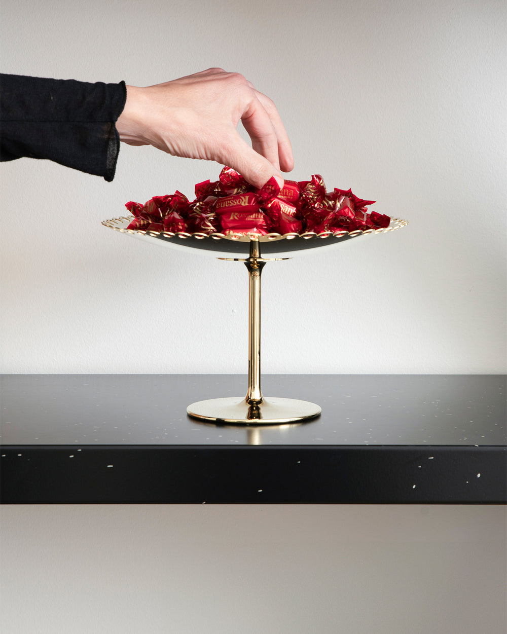 Gold and silver centerpiece stand by Alessi with a scalloped edge, filled with red-wrapped candies, a hand reaching.