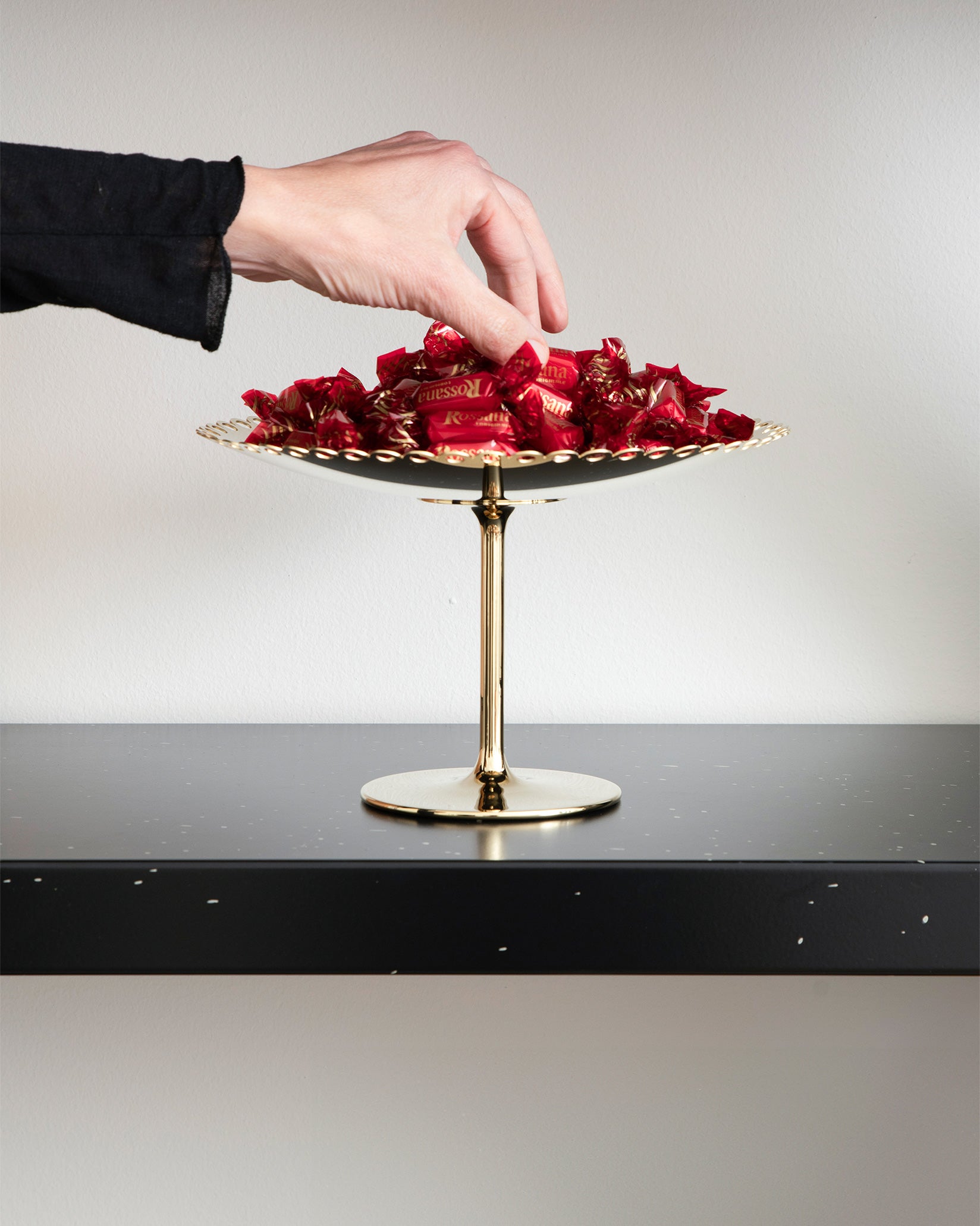 Gold and silver centerpiece stand by Alessi with a scalloped edge, filled with red-wrapped candies, a hand reaching.