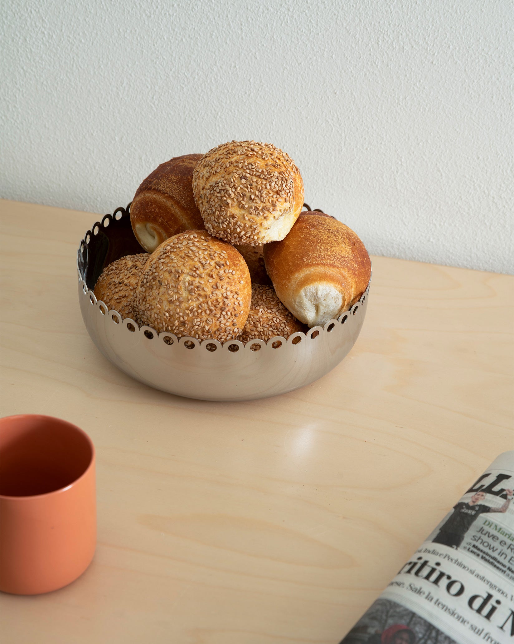 Silver-toned stainless steel bowl by Alessi with a scalloped edge, filled with bread, on a light wood table.