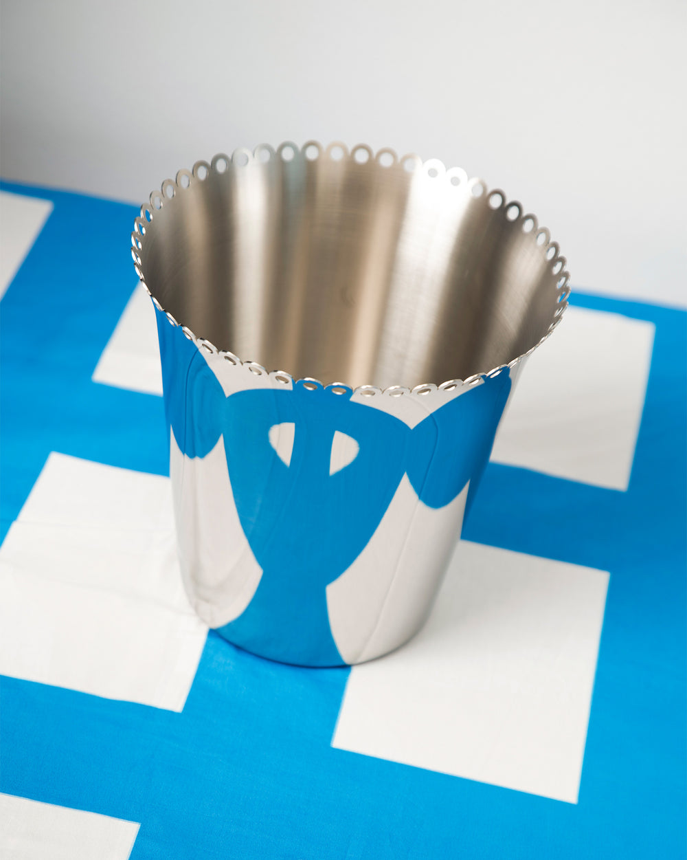 Stainless steel wine cooler by Alessi with a blue and white design and perforated scalloped top edge.