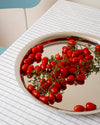 Silver round tray by Alessi filled with red cherry tomatoes, sitting on a white grid-patterned tablecloth.