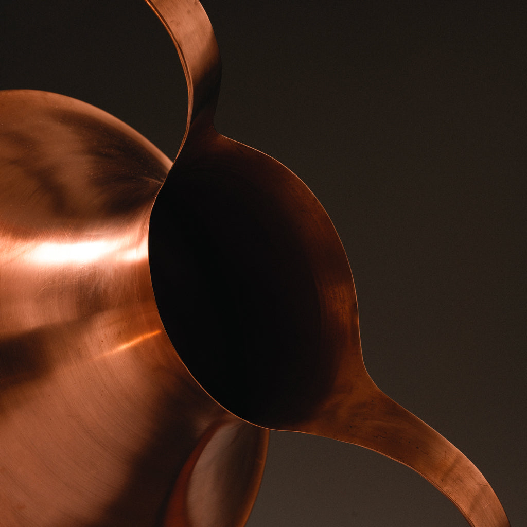 Close-up of a polished copper pitcher with curved spout, warm lighting, dark background.