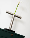 Brushed steel T-shaped vase by Alessi, holding a white calla lily, on a black base above a dark green cabinet.
