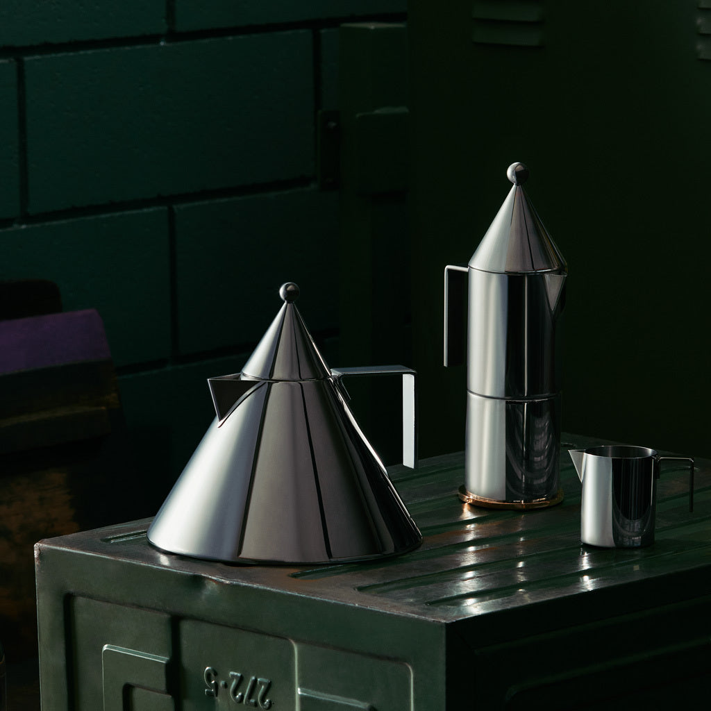 Three sleek, cone-shaped stainless steel coffee makers and a pitcher on a dark green metal chest against a brick wall.