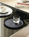 Black and silver pleated metal trays by Alessi, holding coffee and water on a reflective table.