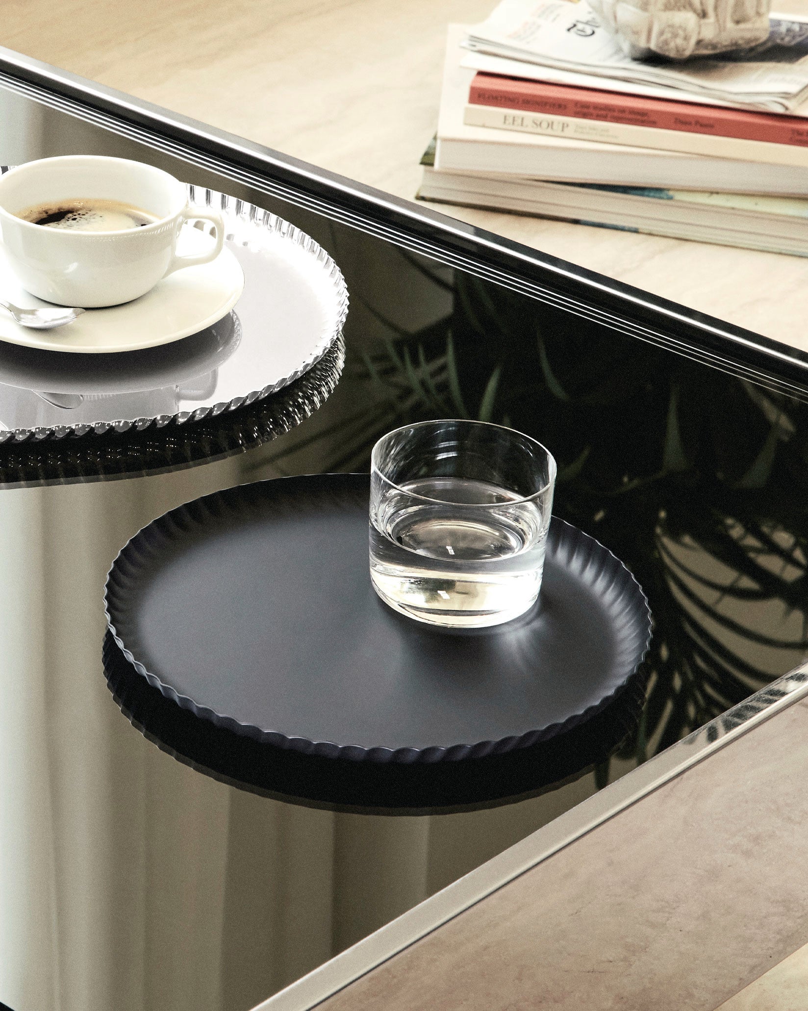 Black and silver pleated metal trays by Alessi, holding coffee and water on a reflective table.