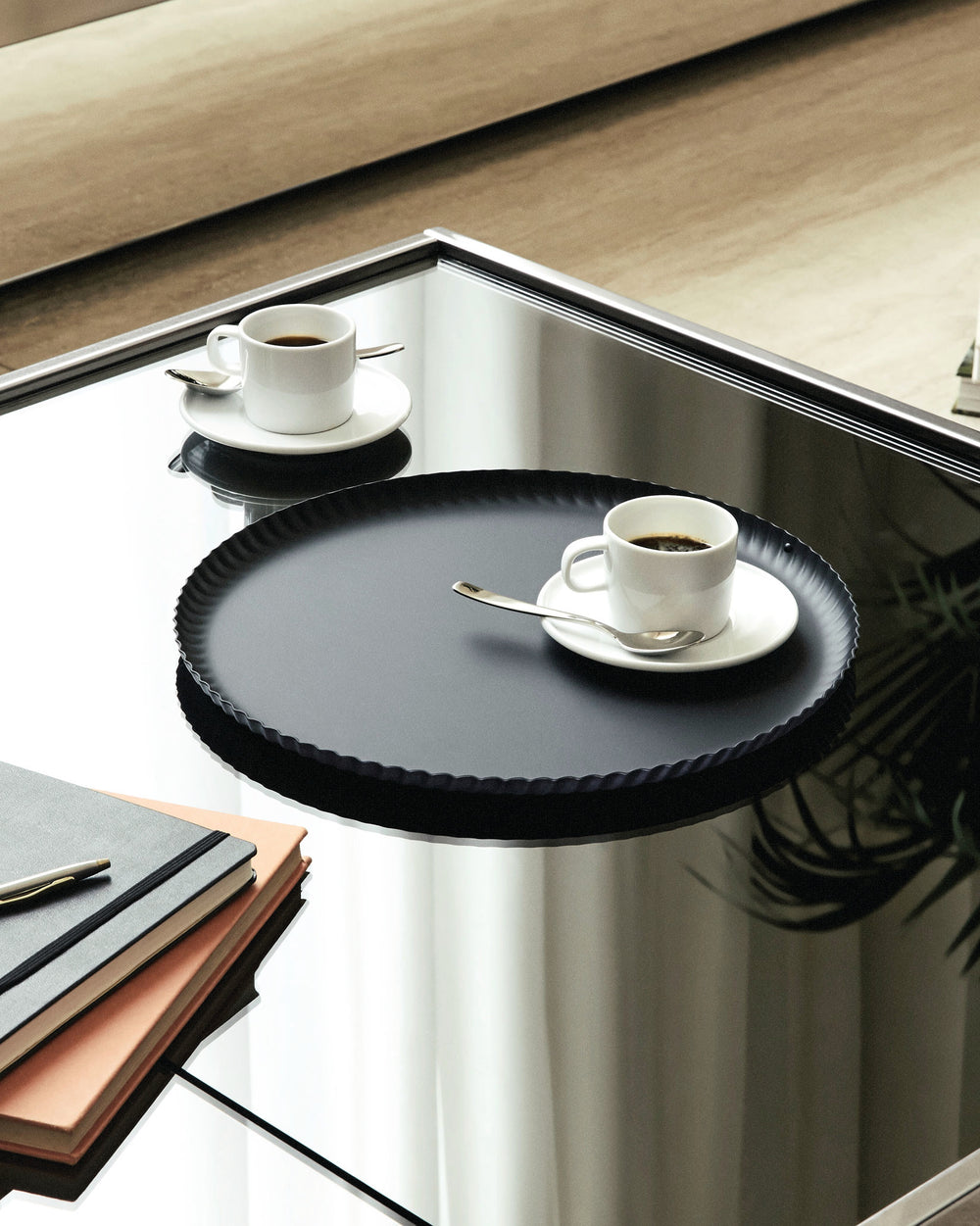 Black tray with a ruffled edge by Alessi, presented on a reflective coffee table with two coffee cups and notebooks.