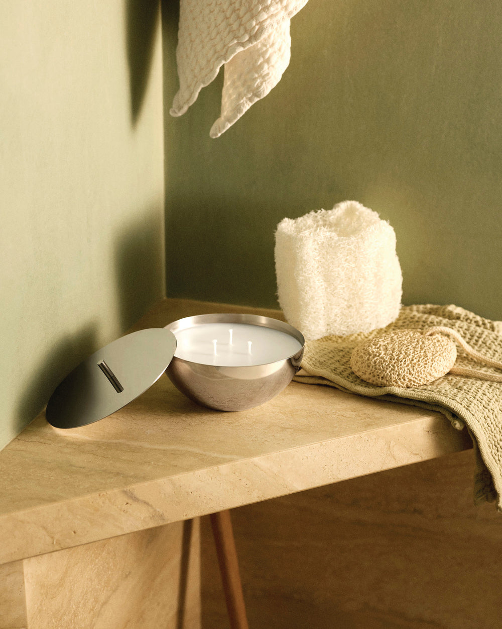 Silver multi-wick candle by Alessi in a polished bowl with its matching lid, on a light brown counter.