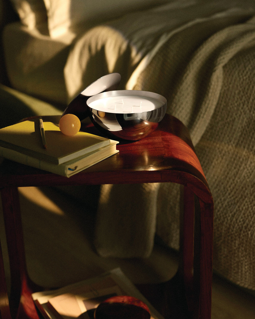 White candle in a polished silver bowl by Alessi, placed on a wooden side table with books and a yellow sphere.