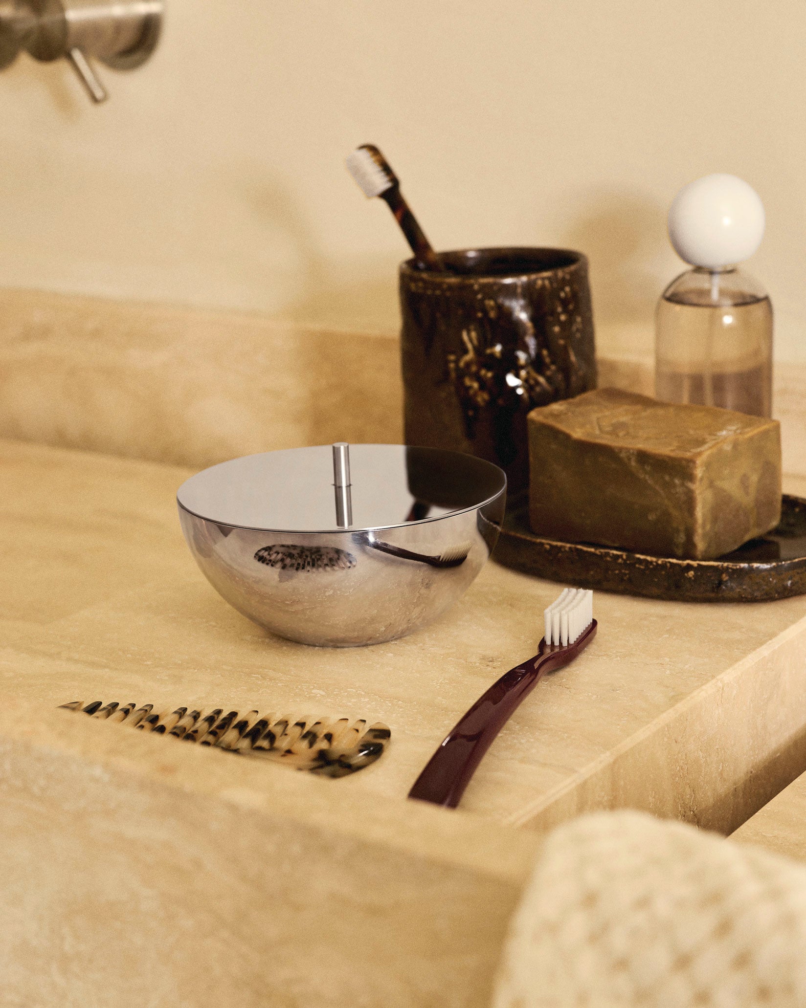 Polished stainless steel lidded container by Alessi, with a cylindrical handle on top, reflecting bathroom items on a beige countertop.