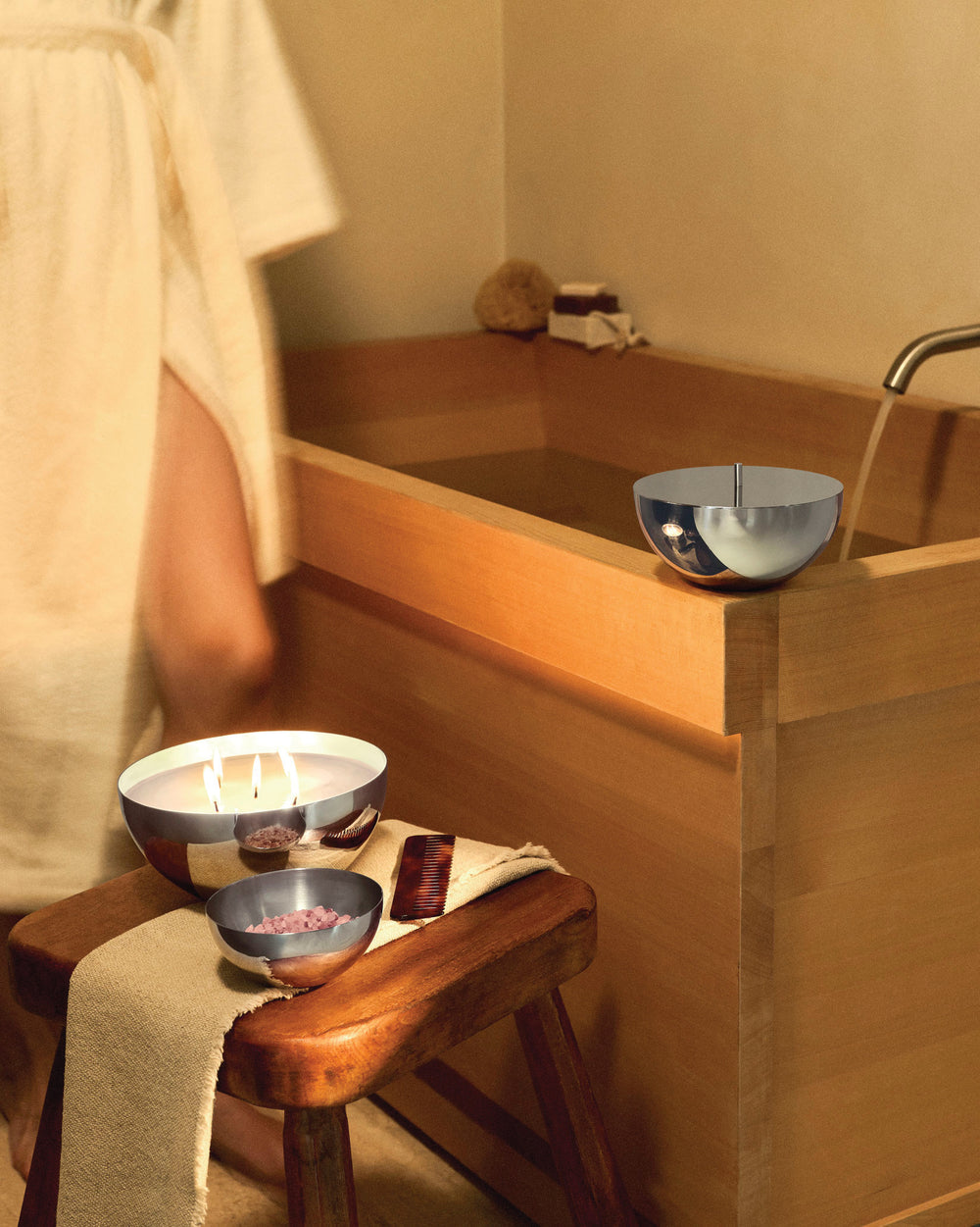 Three-wick candle in a silver-toned bowl by Alessi, lit on a wooden stool with bath salts and a comb next to a wooden tub.