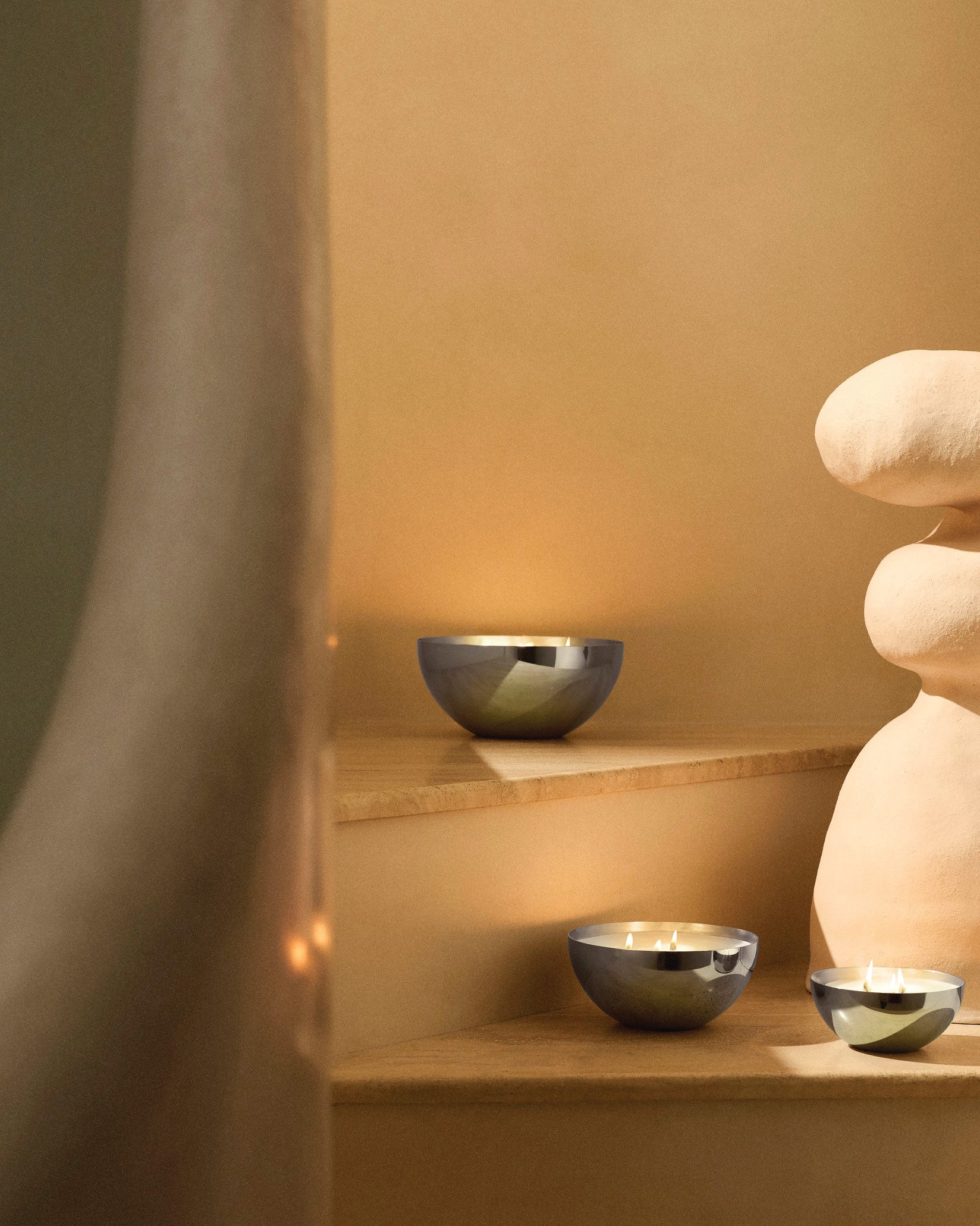 Three metallic bowl-shaped candles by Alessi with glowing wicks on warm steps next to a sculptural piece.