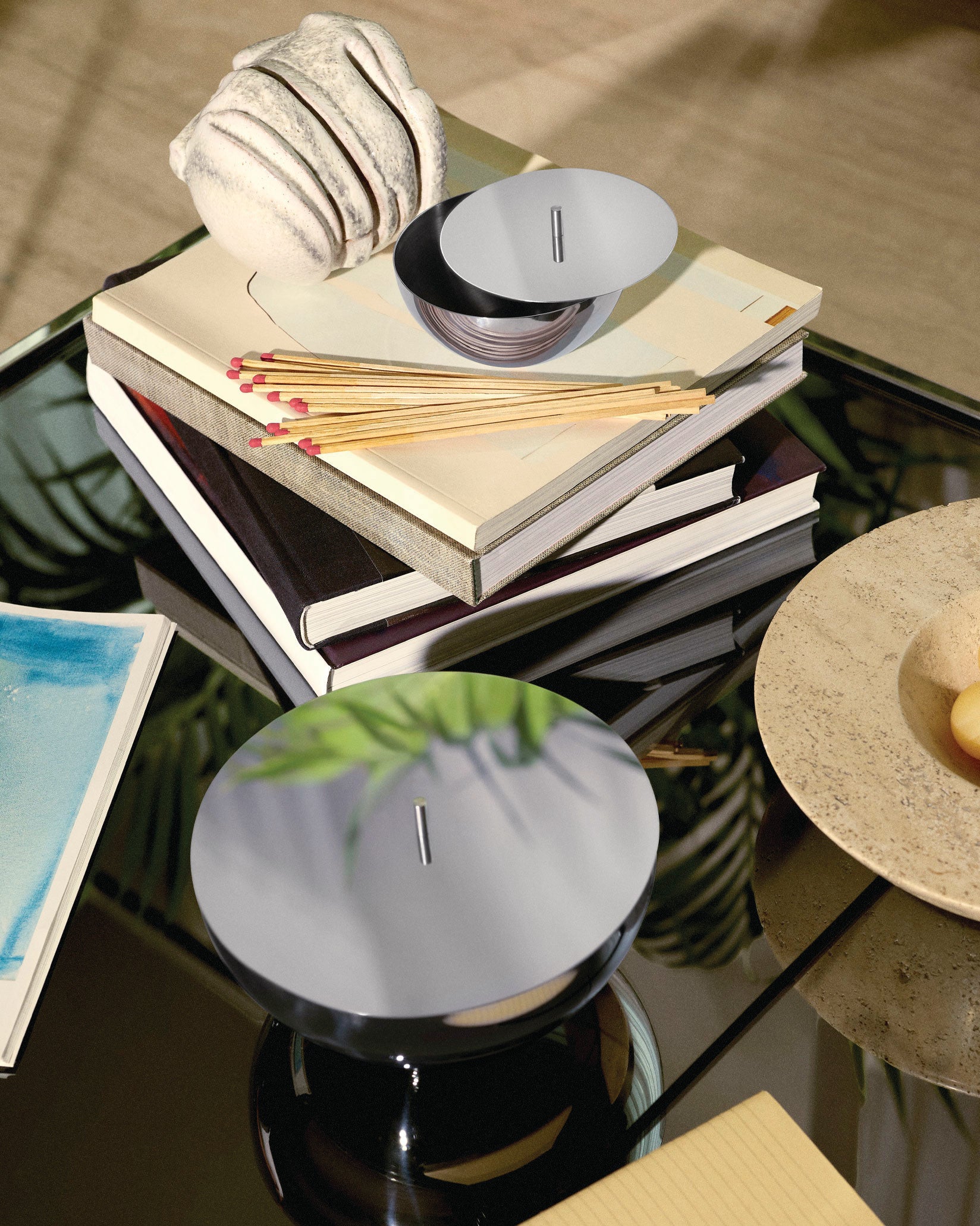 Polished chrome Rocc container by Alessi, with a round flat lid, styled on a glass table with books.