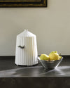 White pleated ceramic water dispenser with conical lid and chrome spout, displayed on dark countertop beside metallic bowl of lemons.