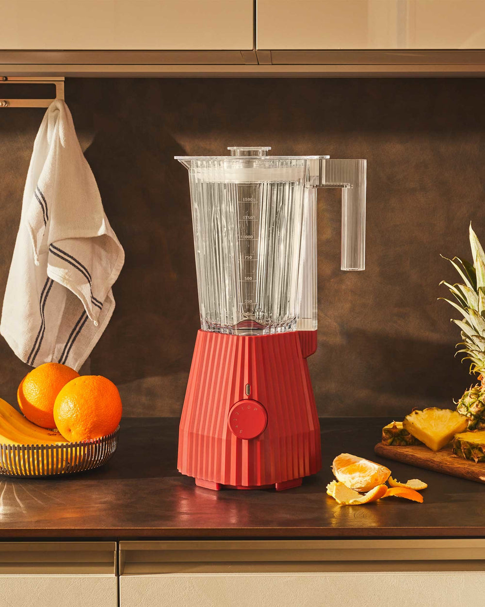 Red pleated blender by Alessi with a clear pitcher and control dial, on a kitchen counter with fruit.