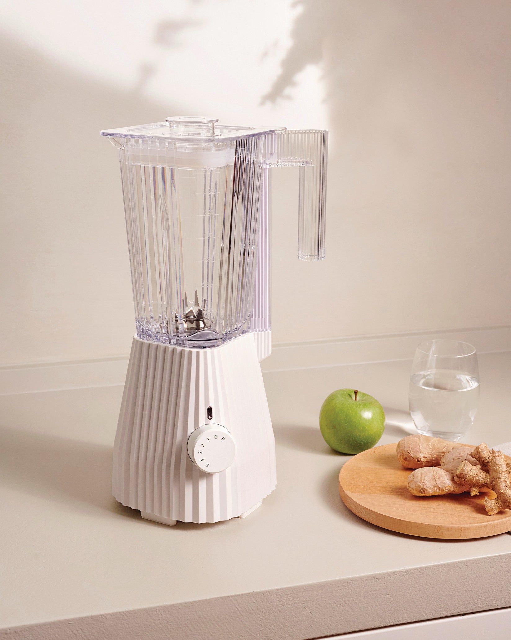 White pleated blender by Alessi with a clear pitcher, control dial, styled on a counter with an apple, water, and ginger.