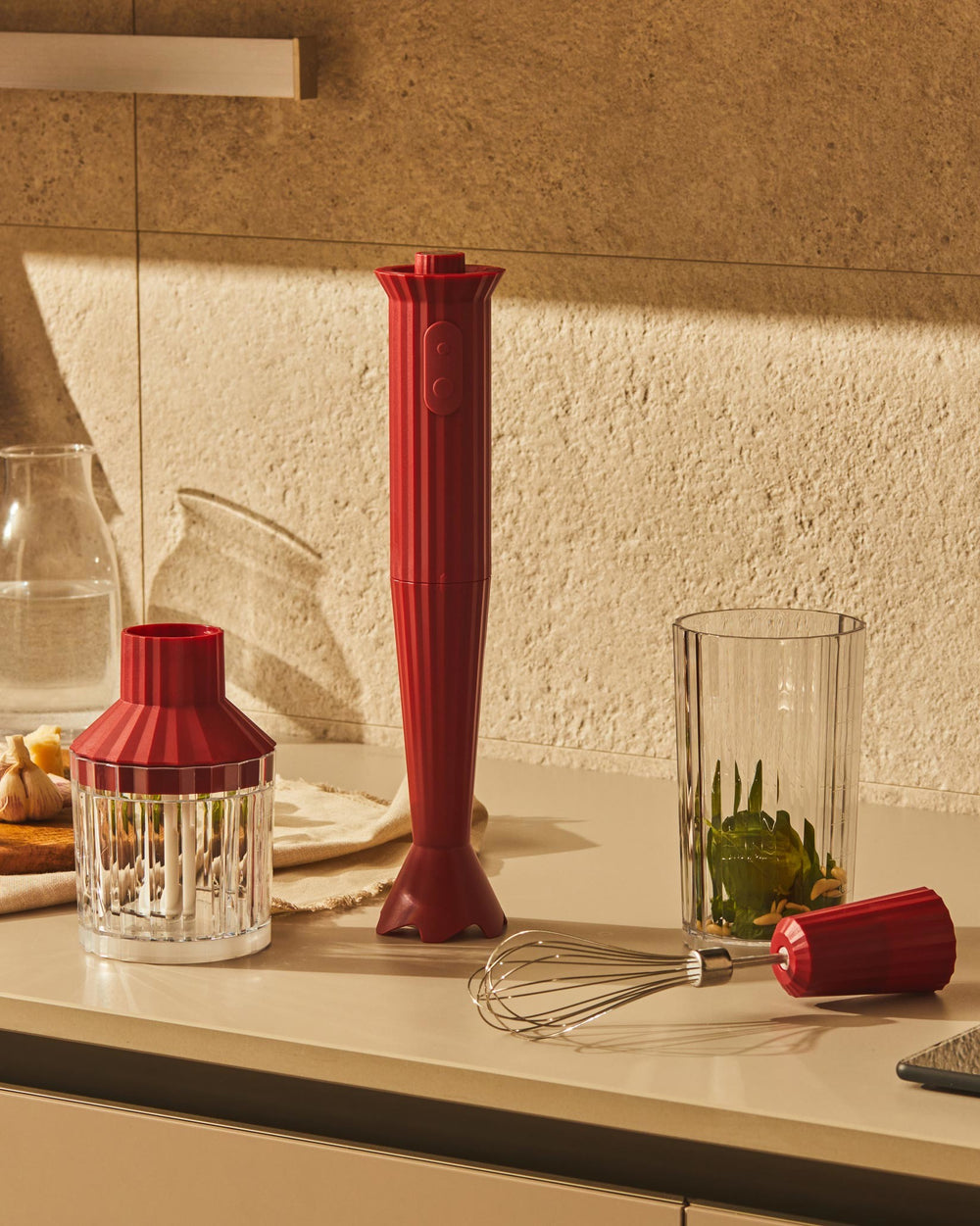 Red ribbed hand blender by Alessi, standing upright with chopper lid, whisk, and pesto ingredients in a glass.