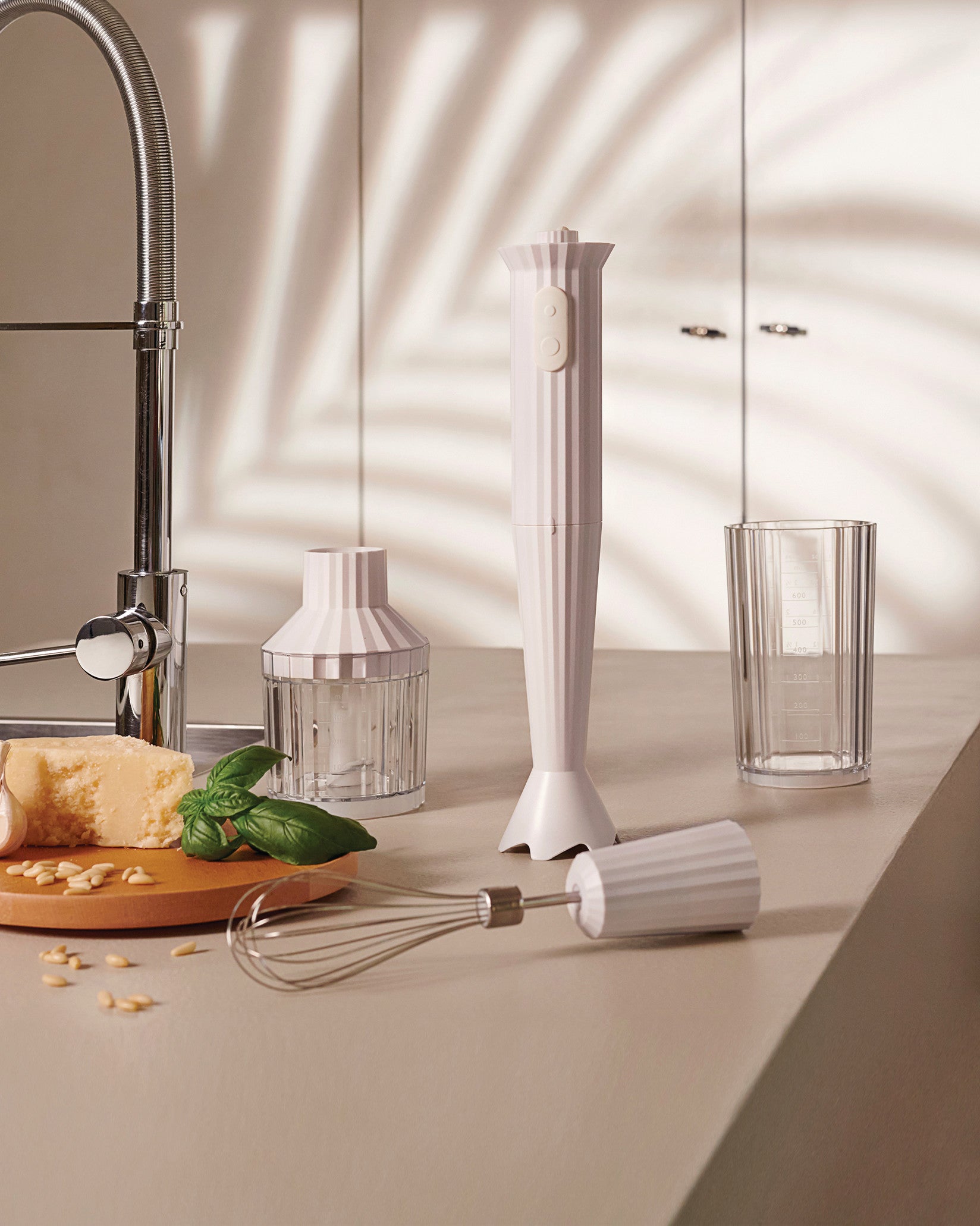 White pleated hand blender by Alessi with matching whisk, chopper, and measuring cup on a kitchen counter.