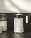 White ribbed milk skimmer by Alessi with a polished silver pitcher, power button, and control dial, beside a milk carafe.