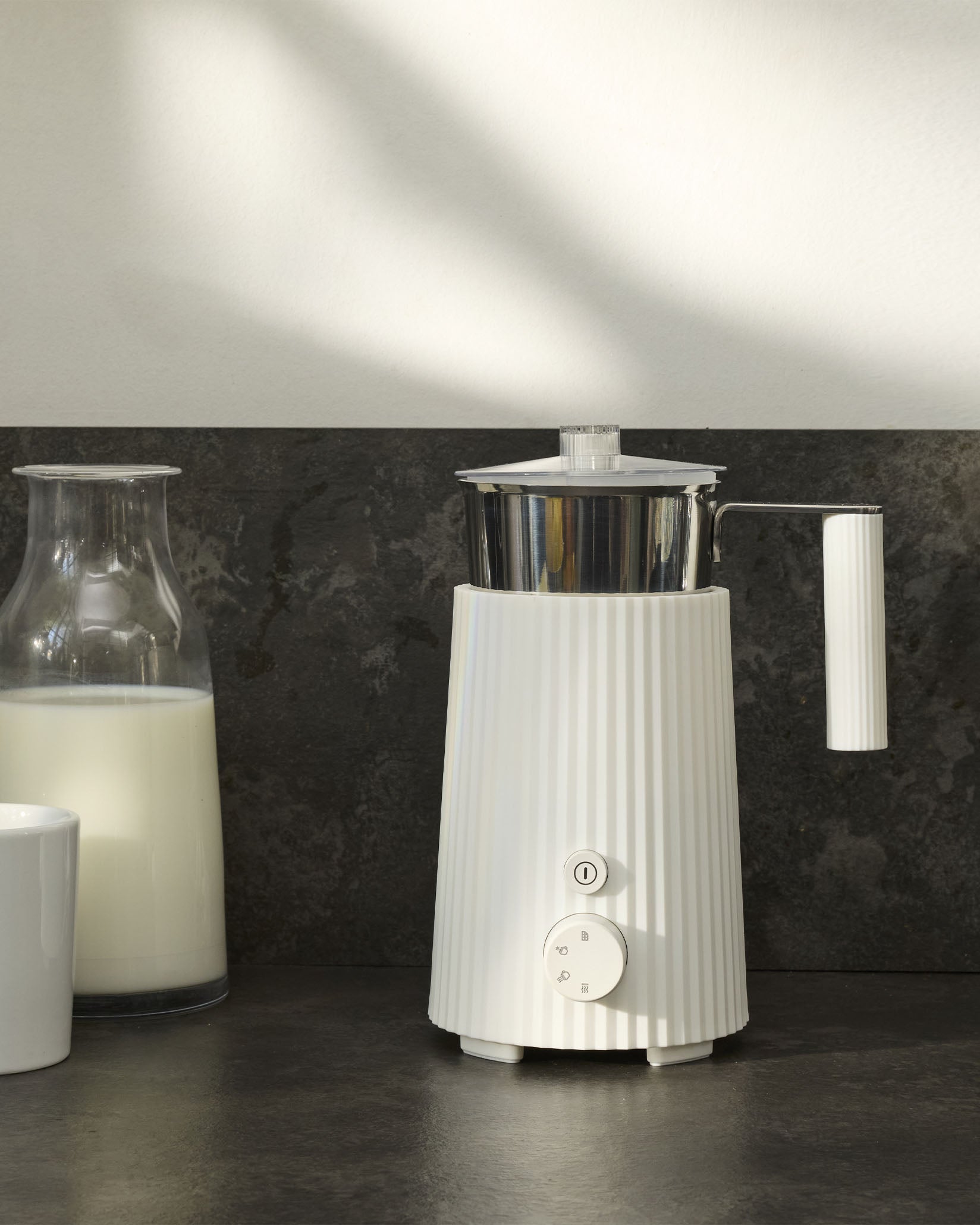 White ribbed milk skimmer by Alessi with a polished silver pitcher, power button, and control dial, beside a milk carafe.