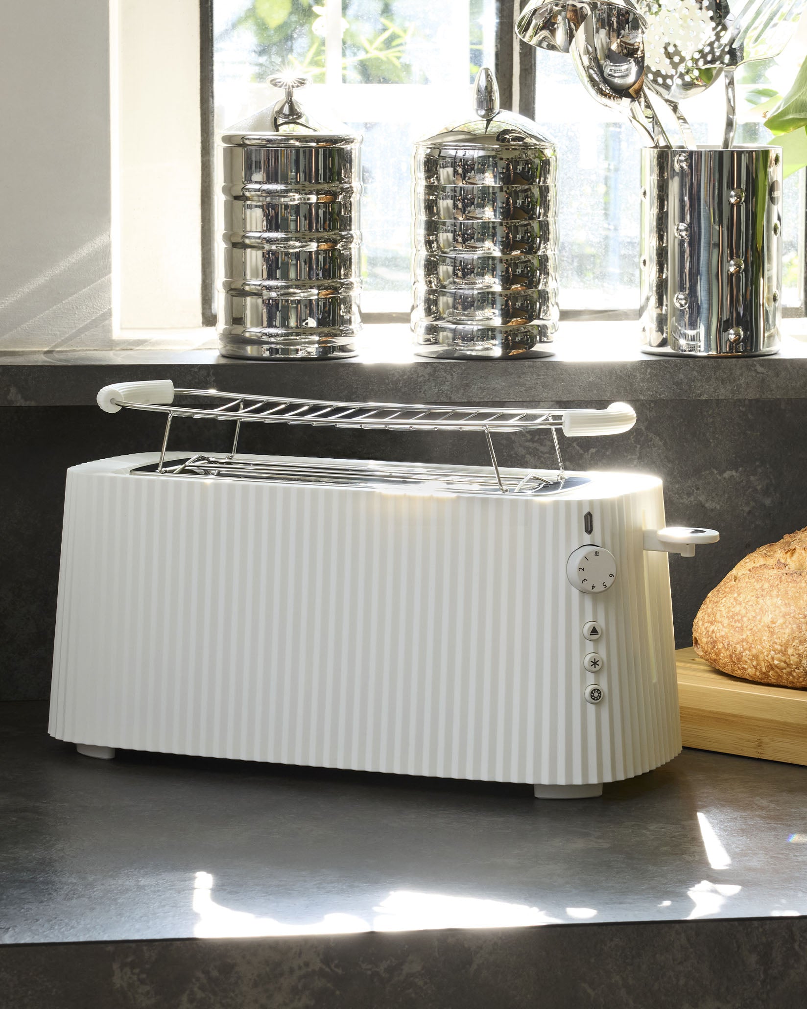 White pleated toaster by Alessi with a chrome warming rack, browning dial, and function buttons on a dark countertop.