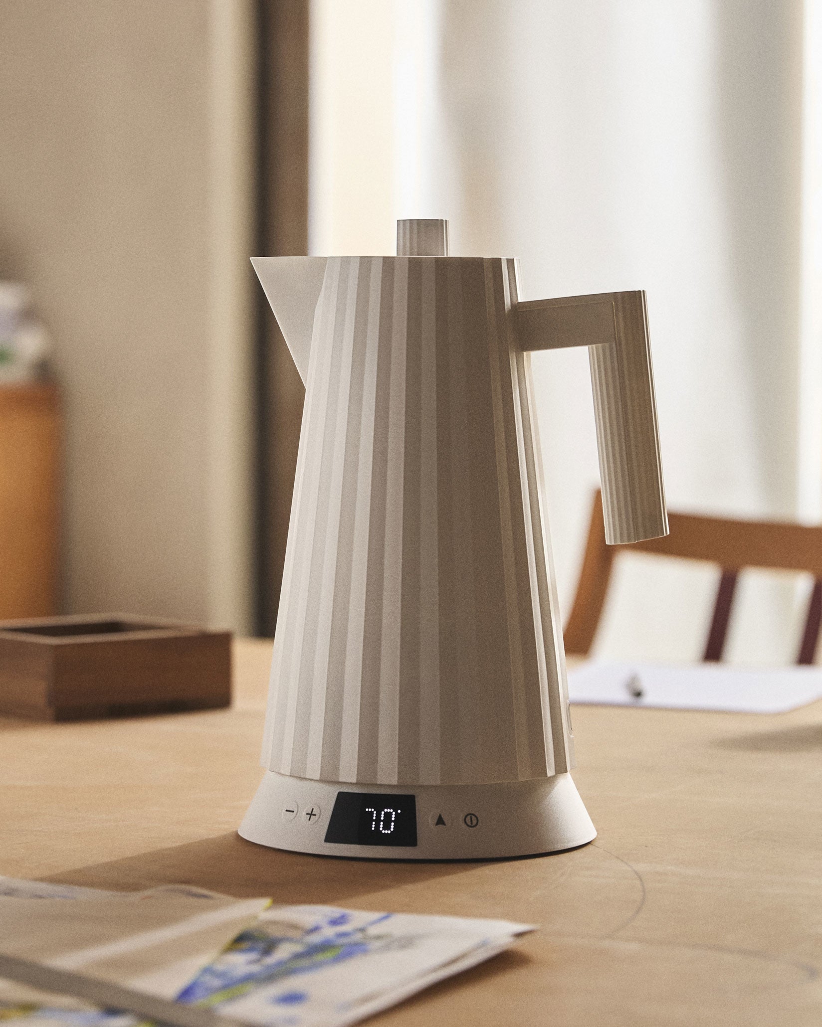 Light grey pleated electric kettle by Alessi with a digital display showing 70° on a wooden table.