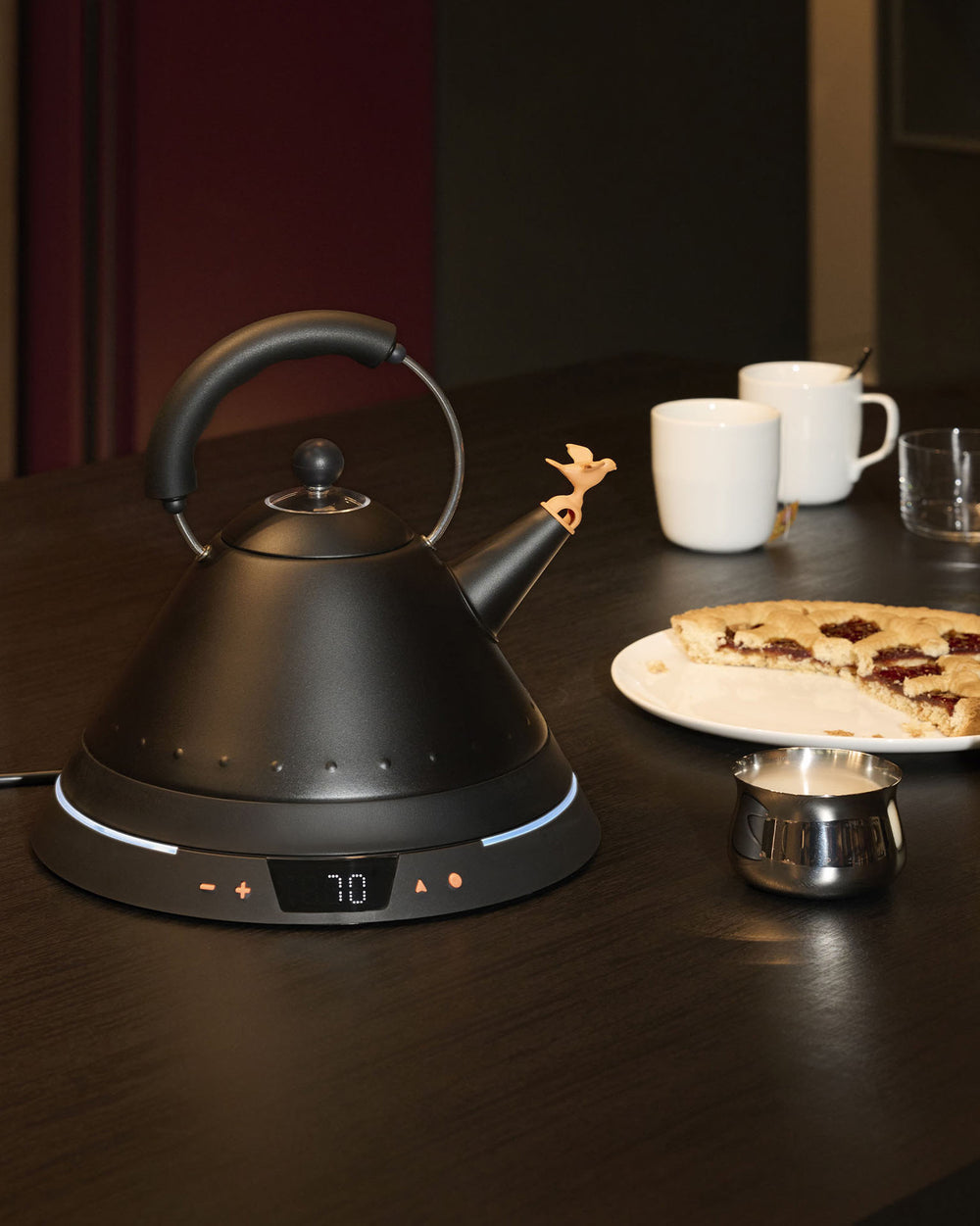 Matte black electric kettle by Alessi with a whimsical bird stopper on its spout and an illuminated digital temperature display.