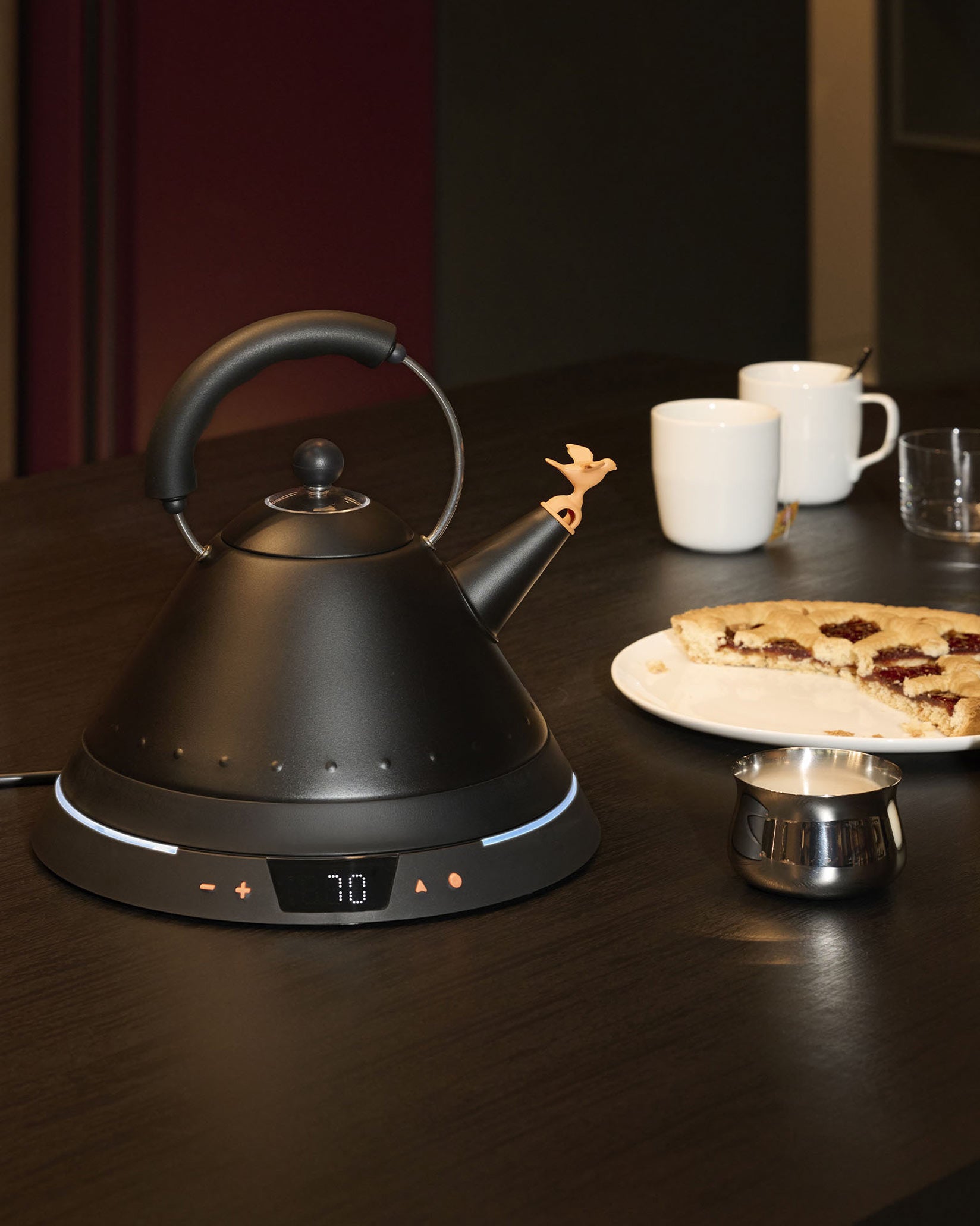 Matte black electric kettle by Alessi with a whimsical bird stopper on its spout and an illuminated digital temperature display.