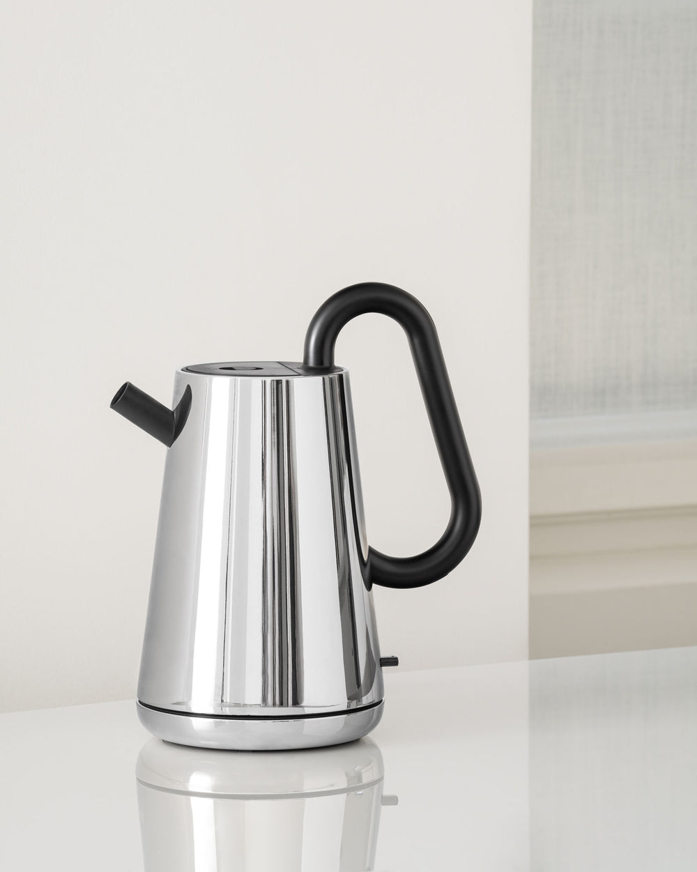 Chrome electric kettle by Alessi with a sleek, modern design, a black looped handle, and a short black spout, on a white reflective surface.