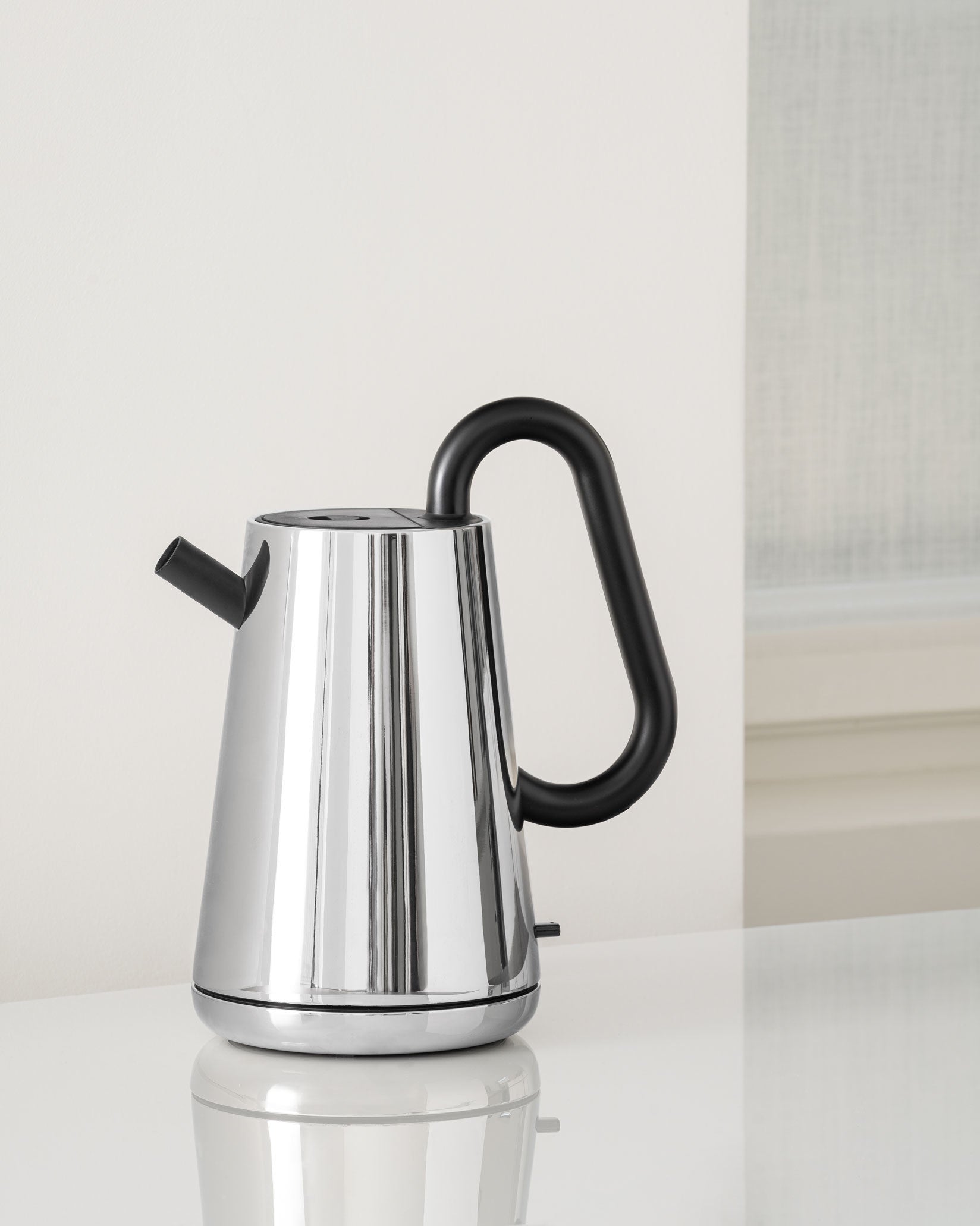 Chrome electric kettle by Alessi with a sleek, modern design, a black looped handle, and a short black spout, on a white reflective surface.