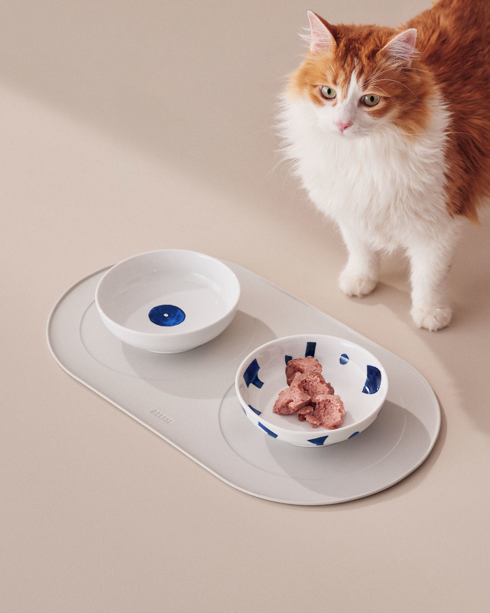 White ceramic cat bowls by Alessi, one with a blue dot, the other with blue shapes and food, on a gray Alessi mat with an orange cat.