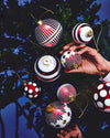 Red and white Christmas tree ornaments by Alessi, featuring striped and polka dot designs, with gold hangers, held by a hand.