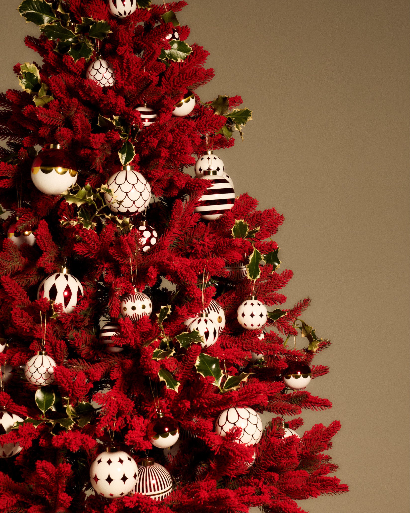 White and red Christmas tree ornaments by Alessi with various patterns, hanging on a deep red tree with green holly leaves.