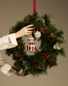 Three white and red geometric Christmas tree ornaments by Alessi on a green wreath, held by hands.
