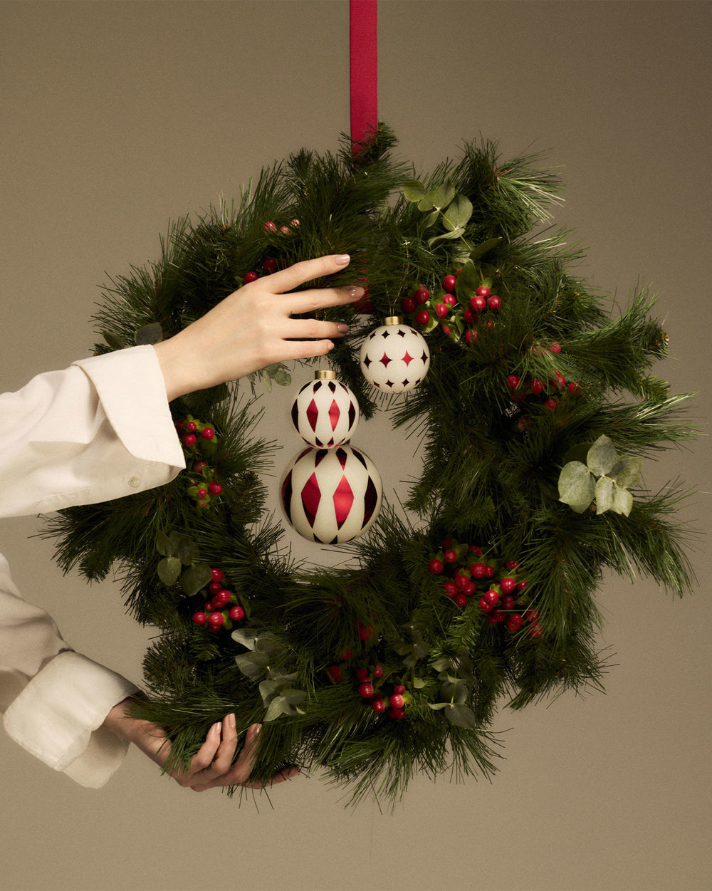 Three white and red geometric Christmas tree ornaments by Alessi on a green wreath, held by hands.
