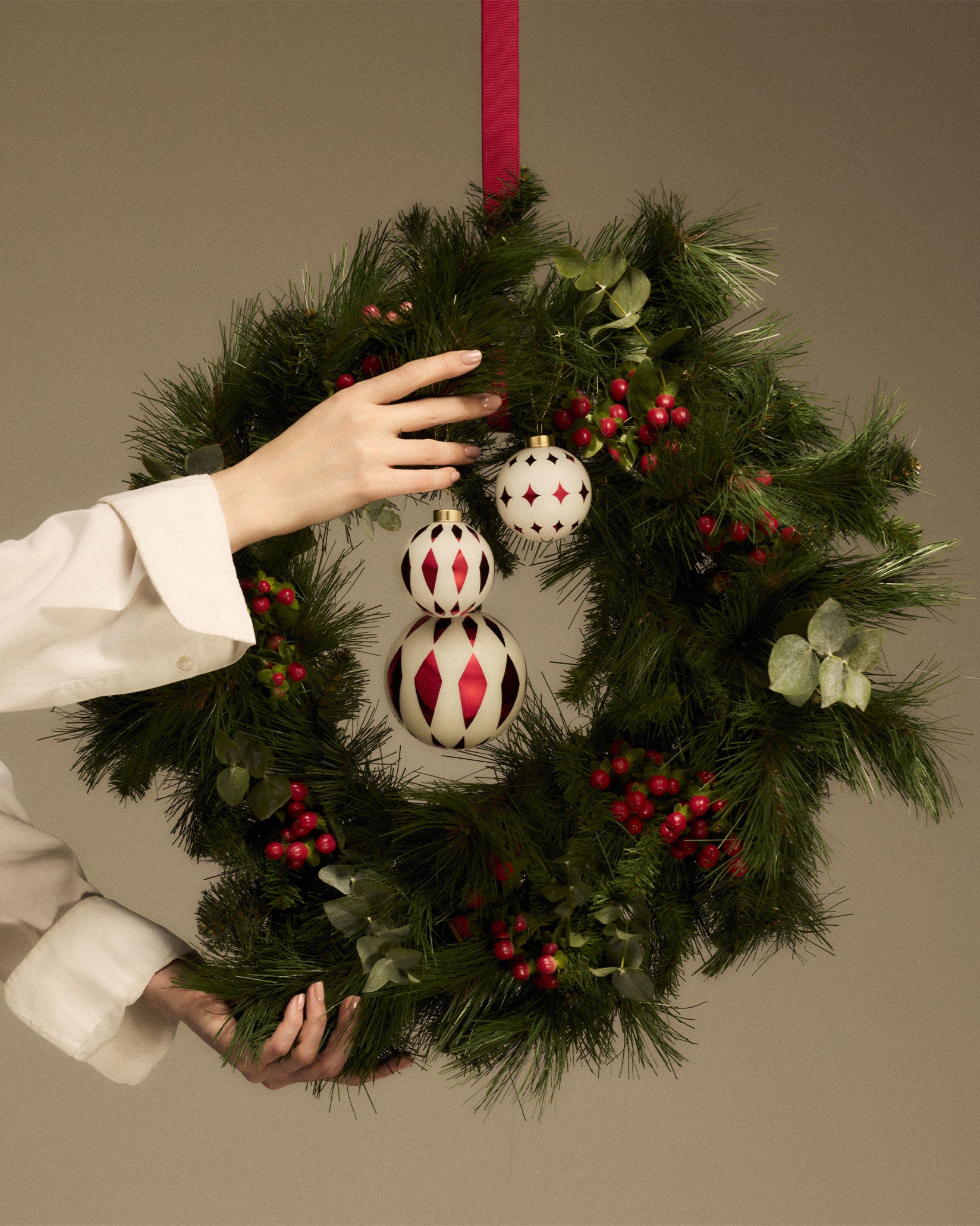 Three white and red geometric Christmas tree ornaments by Alessi on a green wreath, held by hands.