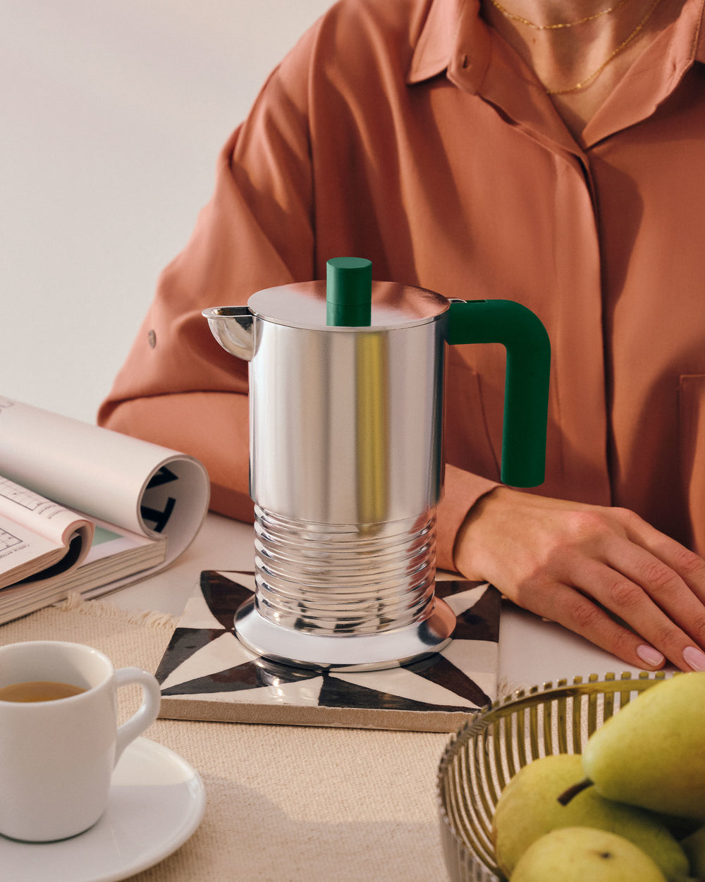 Silver espresso coffee maker with green handle and knob by Alessi, on a patterned coaster next to a coffee cup and pears.